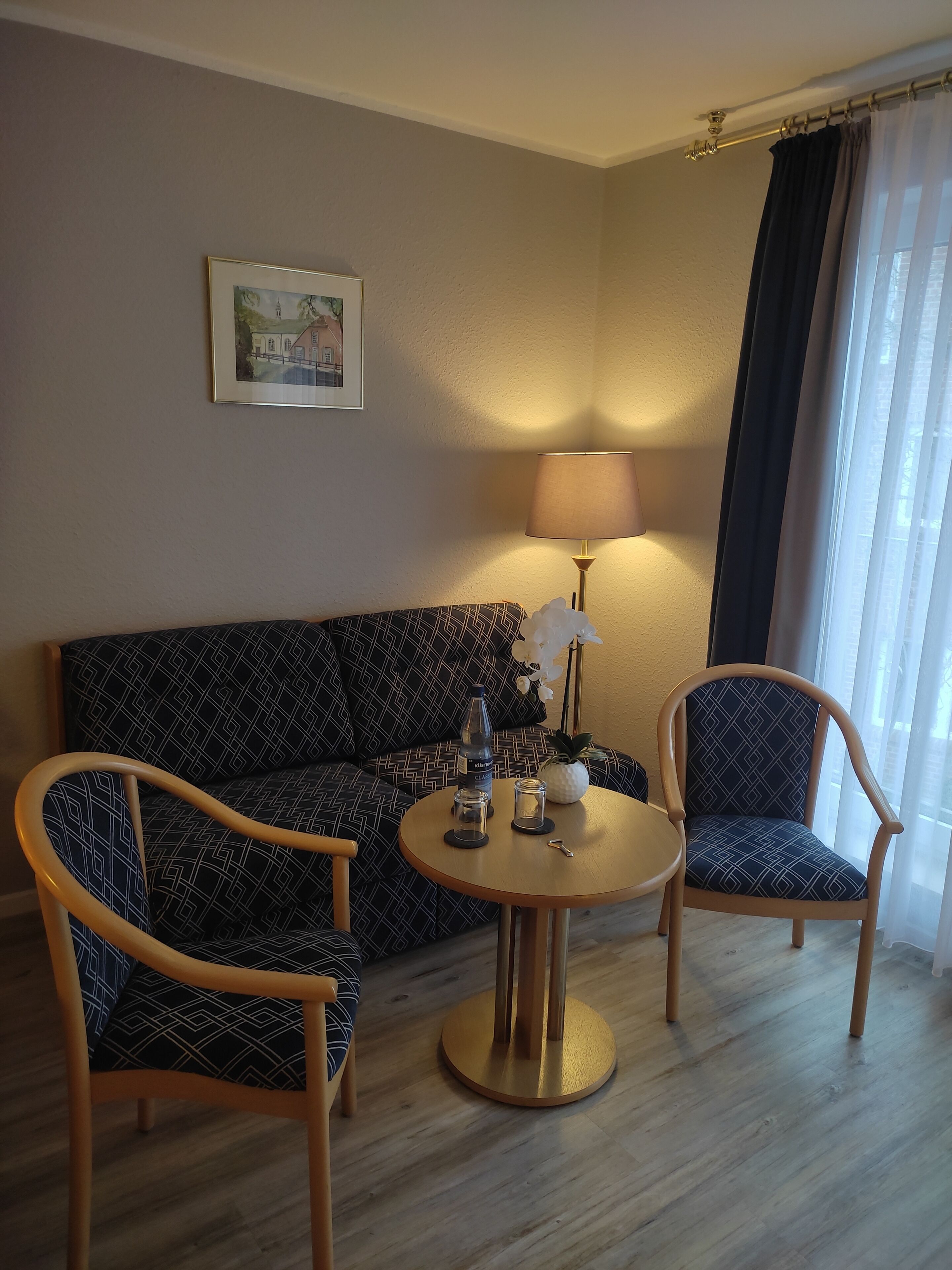 A living room with a sofa, two patterned armchairs, a round coffee table with a bottle, glasses, and a key, a lamp, and a framed picture on the wall.