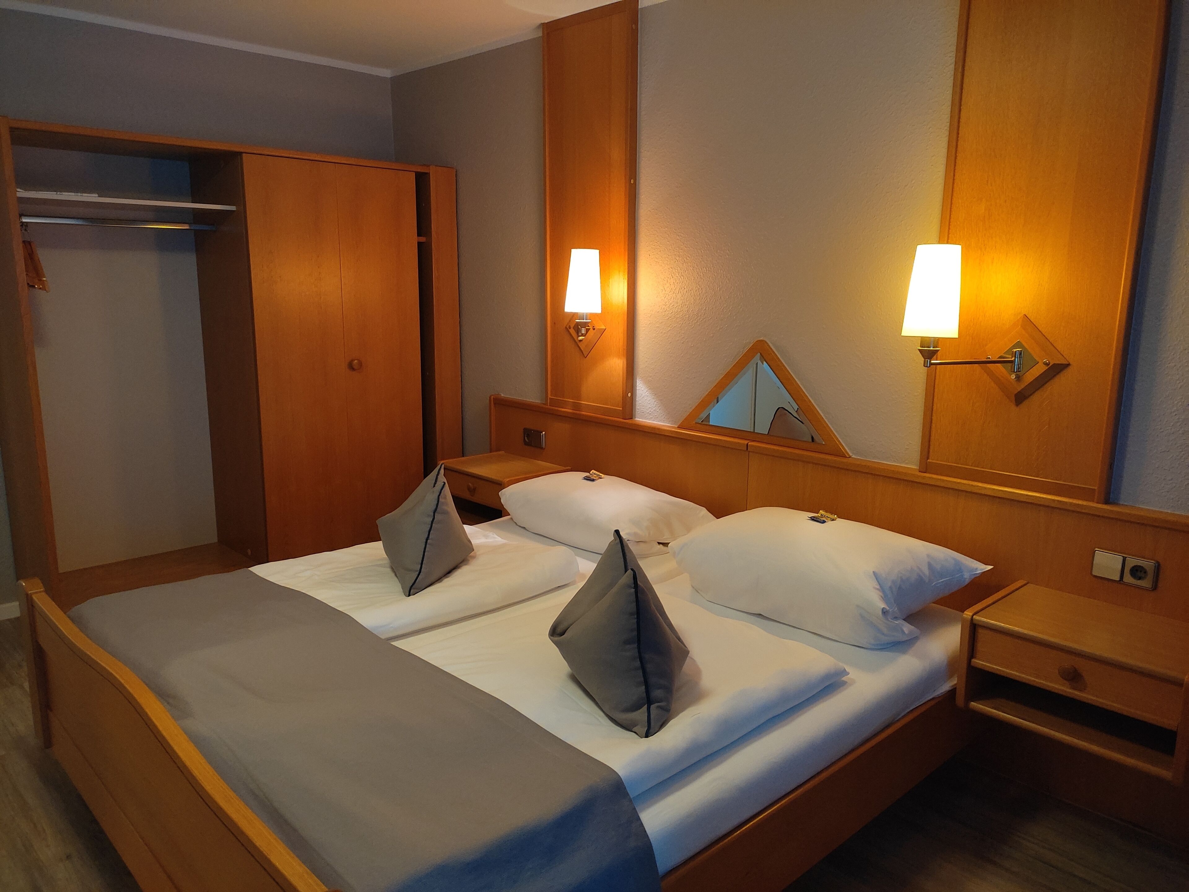 A hotel room with a double bed, wooden headboard, bedside tables, a wardrobe, and wall-mounted lamps.