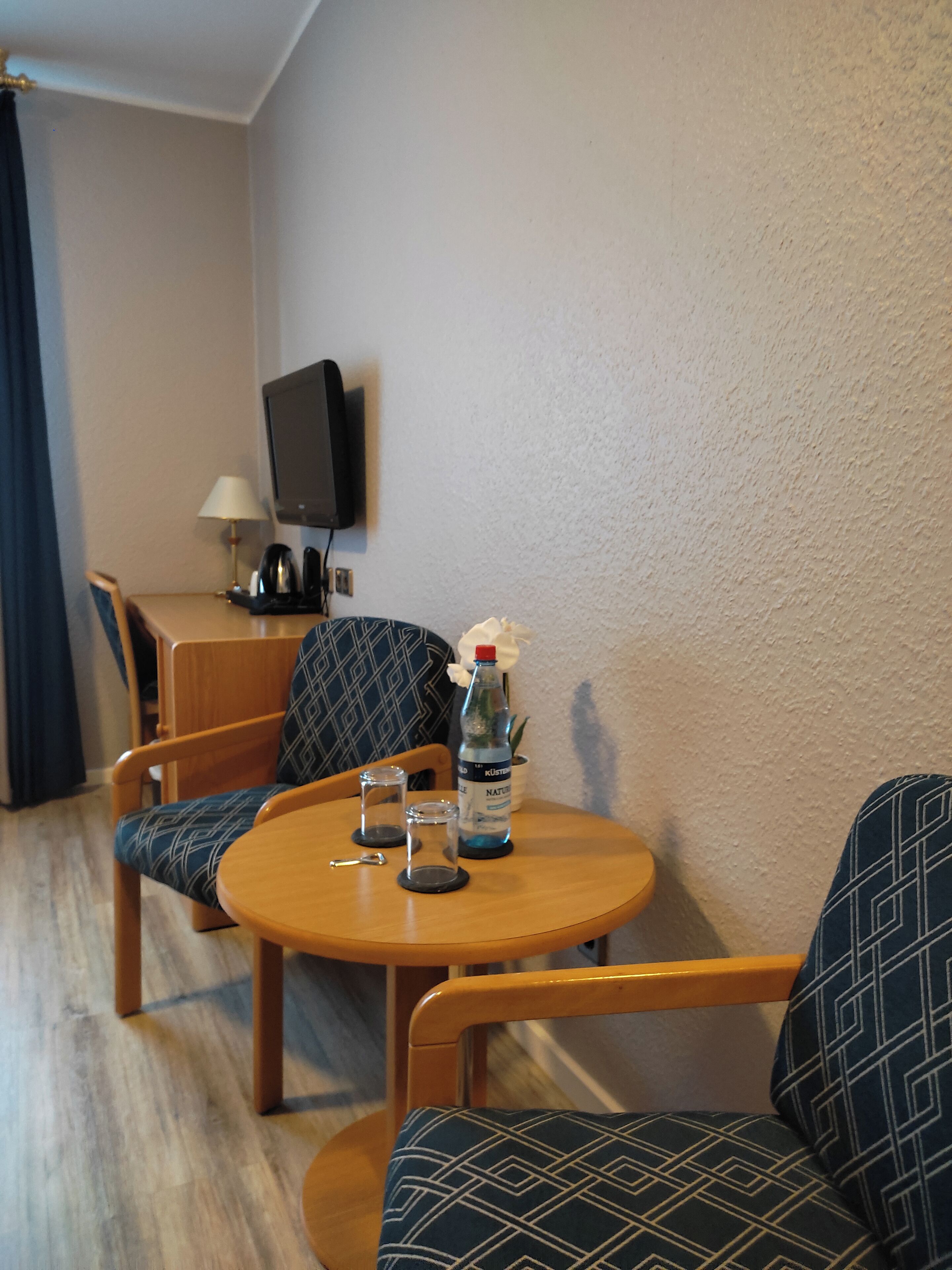 Ein kleiner, gemütlicher Raum mit einem runden Holztisch, zwei gemusterten Stühlen, einem Fernseher an der Wand und einer Wasserflasche.