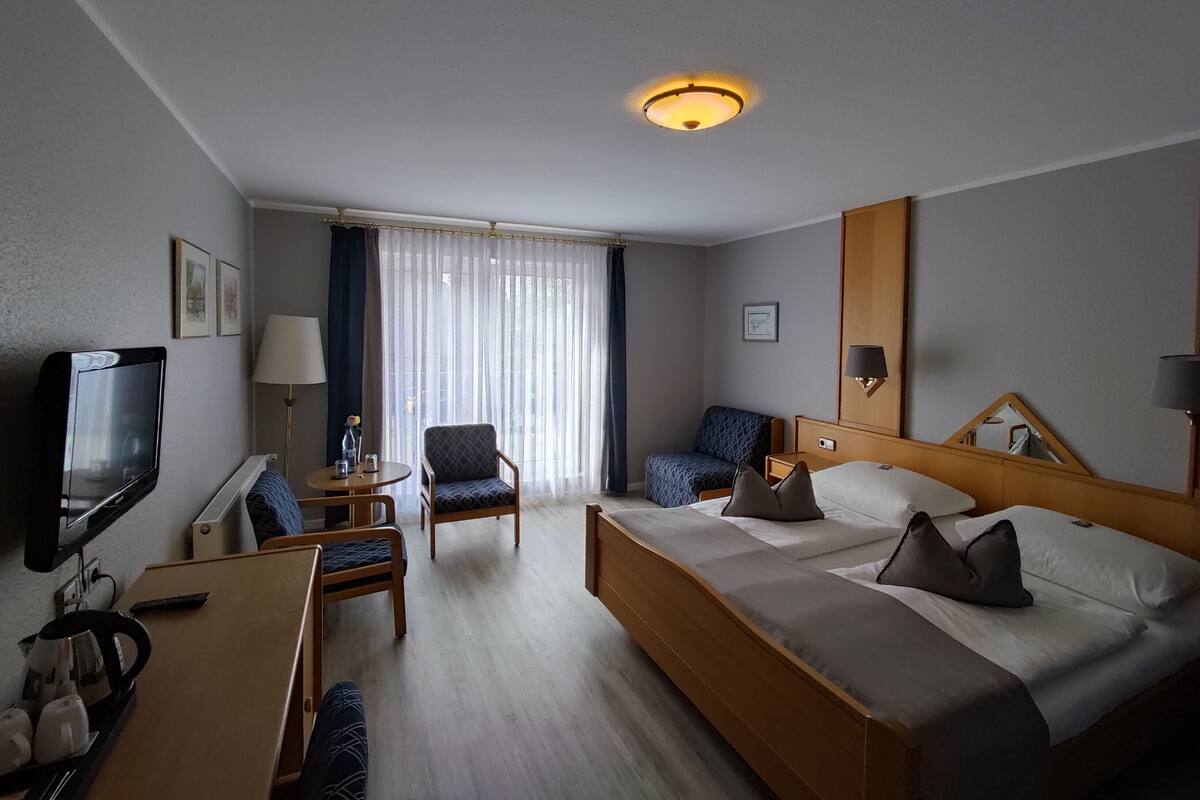 Une chambre d’hôtel comprenant un lit, deux fauteuils, un bureau avec une télévision, une machine à café et une grande fenêtre avec des rideaux.