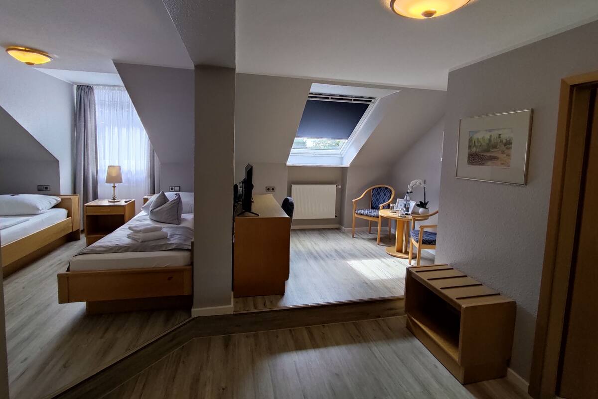 Une chambre d’hôtel moderne au design de type « loft », dotée d’une grande verrière, d’un coin salon confortable avec une table et des chaises, et d’un espace nuit séparé avec un lit et une table de chevet.