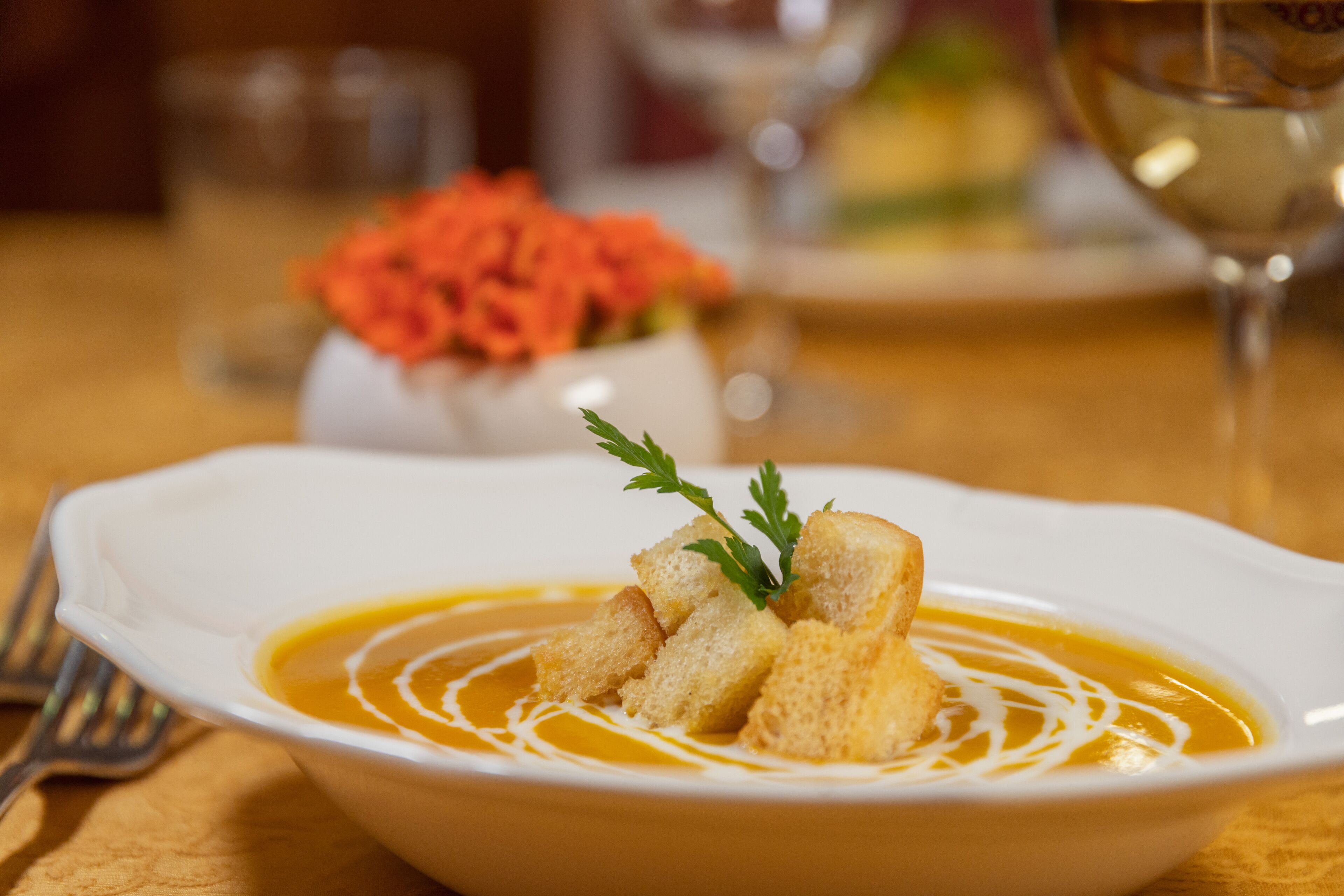Un bol de soupe garni de croûtons et d’herbes, accompagné d’un verre de vin blanc et d’un vase rempli de fleurs oranges.