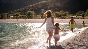A beach scene with a woman and child walking along the shore, a man in the background, and buildings in the distance. - Hotel Miravalle (San Lorenzo Dorsino)