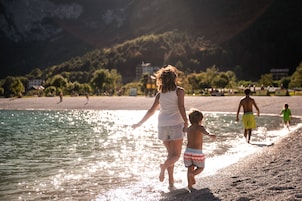 A beach scene with a woman and child walking along the shore, a man in the background, and buildings in the distance.