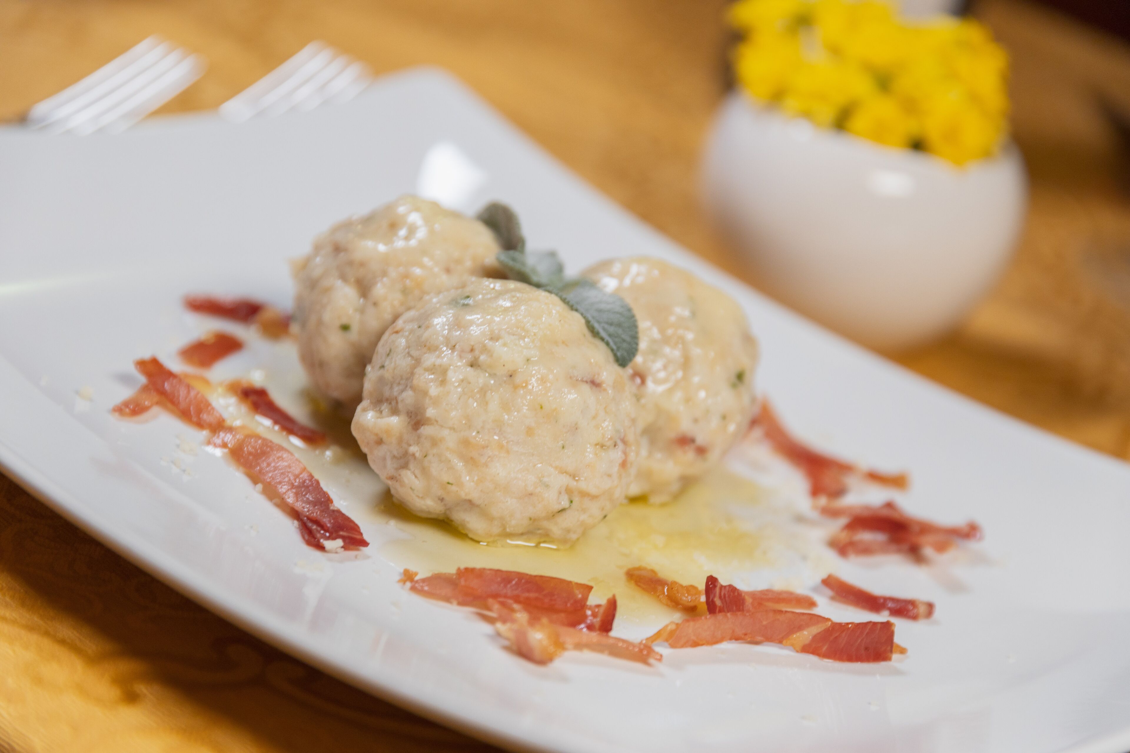 A plate with two meatballs garnished with bacon and a small bowl of yellow flowers in the background.