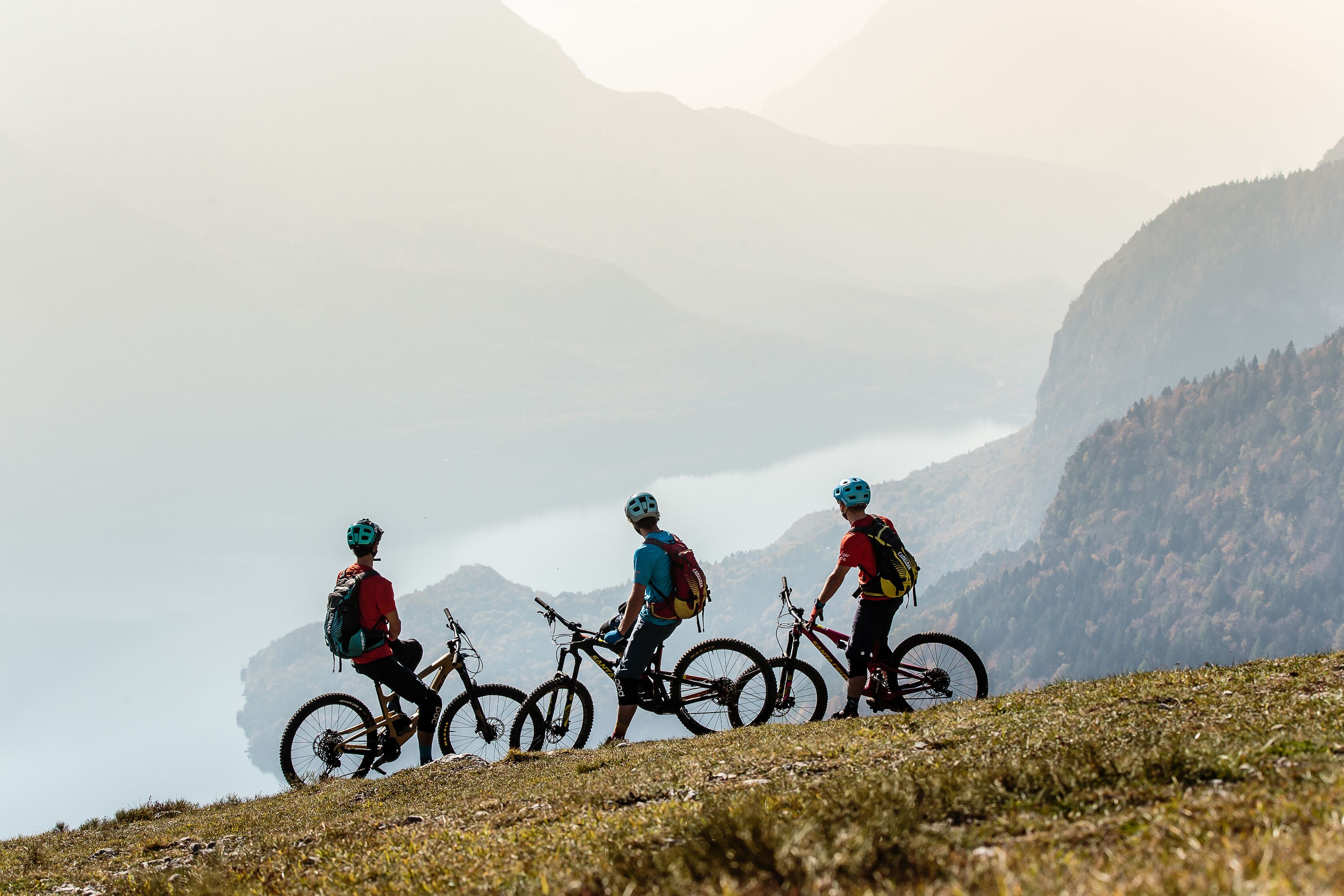 Three mountain bikers on a grassy slope with bicycles, a clear sky, and a mountainous backdrop.