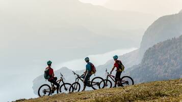 Three mountain bikers on a grassy slope with bicycles, a clear sky, and a mountainous backdrop.