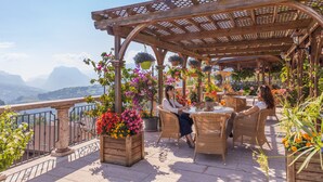 A terrace with wicker furniture, potted plants, and a view of mountains. - Hotel Miravalle (San Lorenzo Dorsino)