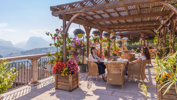 A terrace with wicker furniture, potted plants, and a view of mountains. - Hotel Miravalle (San Lorenzo Dorsino)