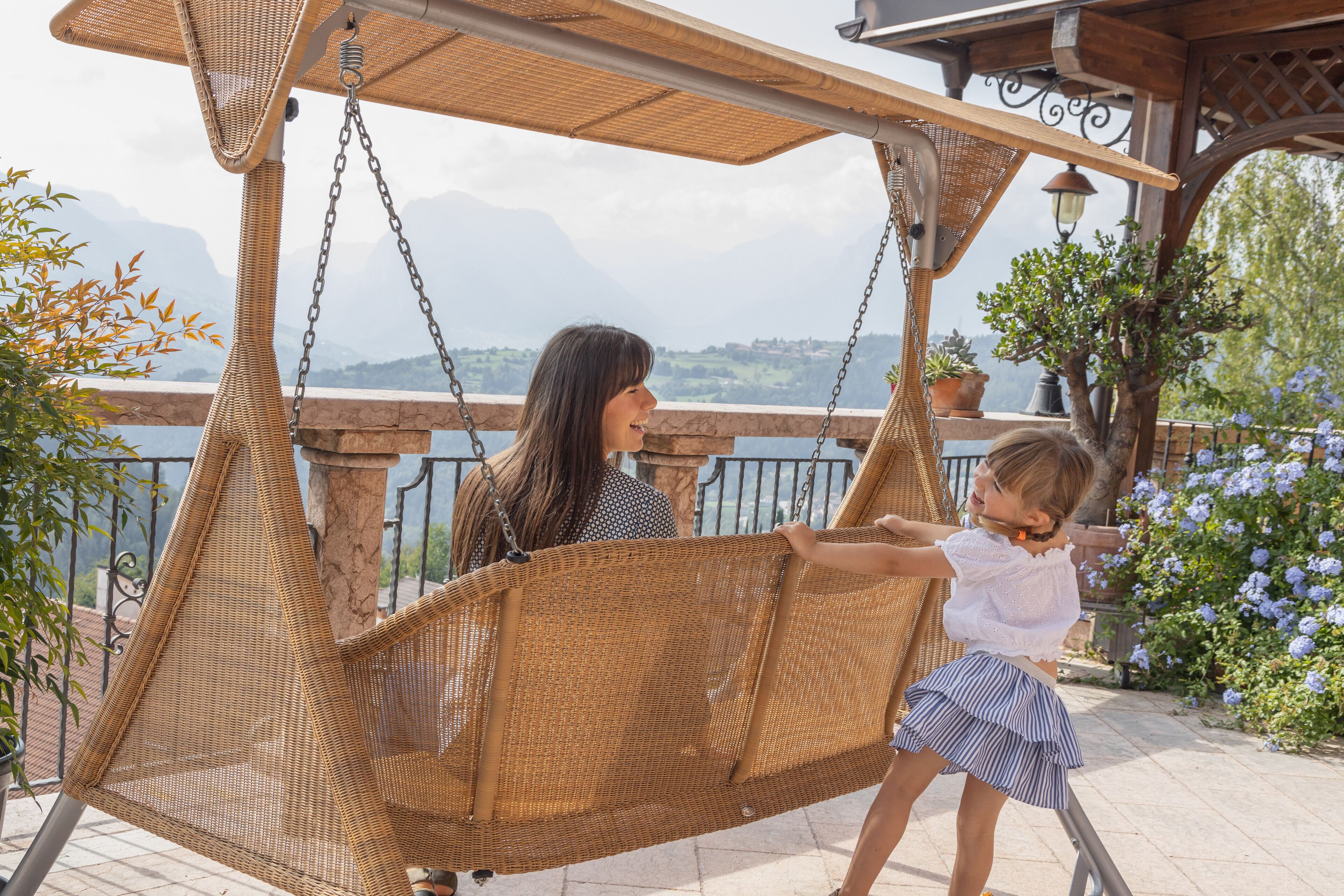 Un fauteuil suspendu en osier, fixé à une pergola, offrant une vue sur les montagnes et un jardin fleuri.