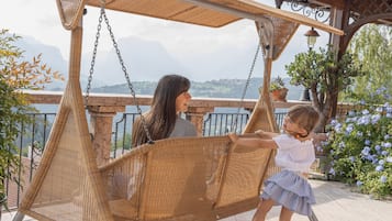 Un fauteuil suspendu en osier, fixé à une pergola, offrant une vue sur les montagnes et un jardin fleuri.