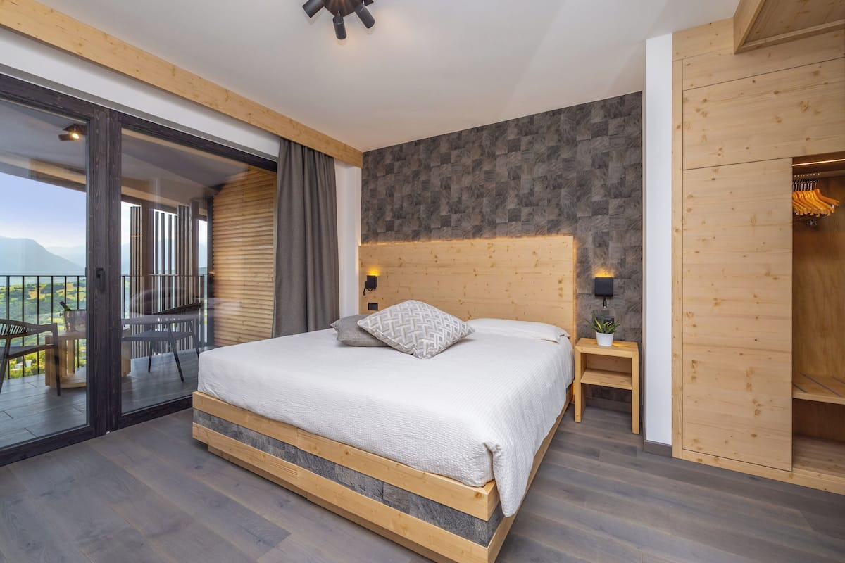 A modern bedroom with a large bed, wooden headboard, and a view of mountains through the balcony.