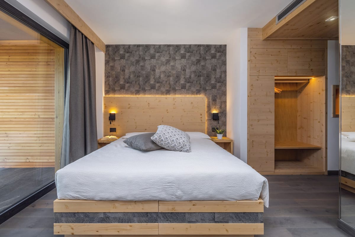A modern bedroom with a wooden headboard, a large bed with a white bedspread, two bedside tables with lamps, and a wooden wardrobe.