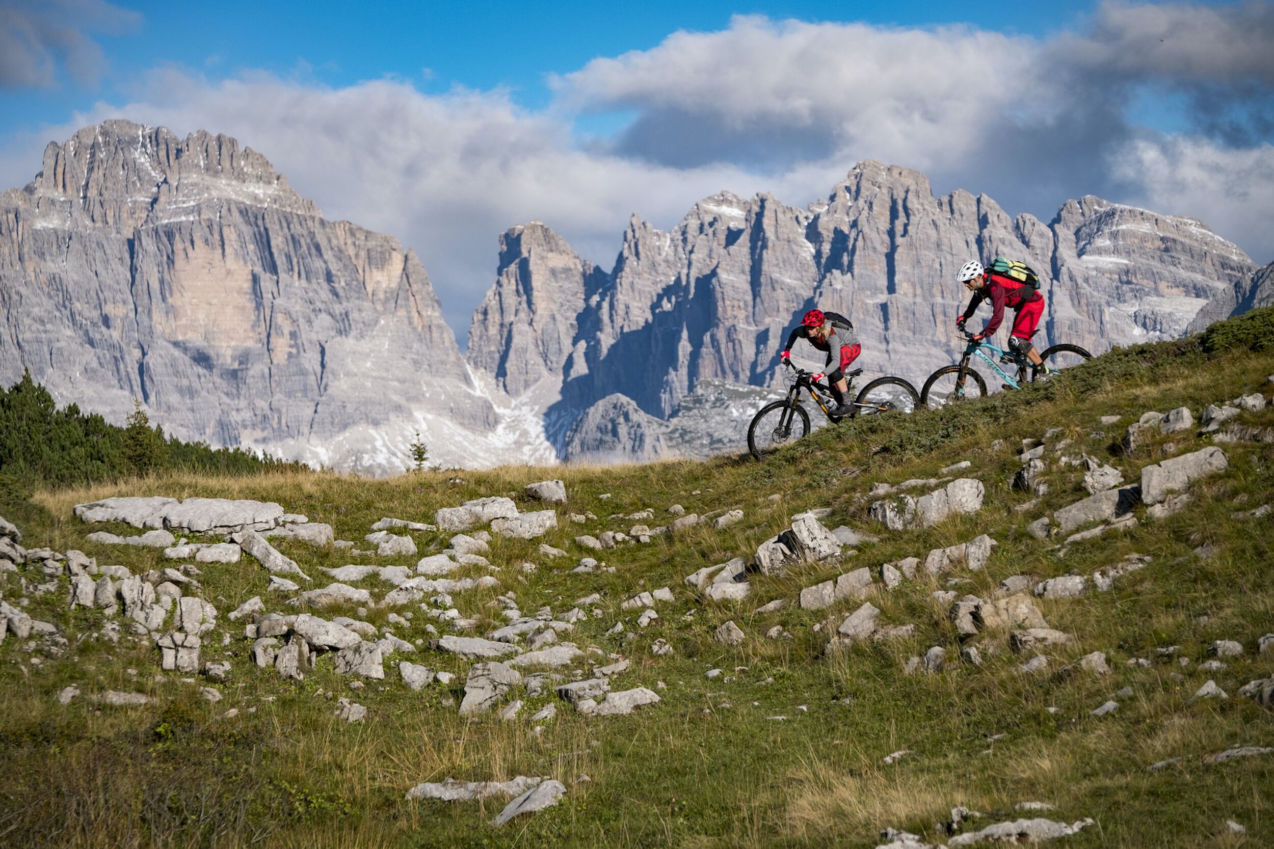 Two cyclists on a rocky mountain path with a mountainous backdrop.
