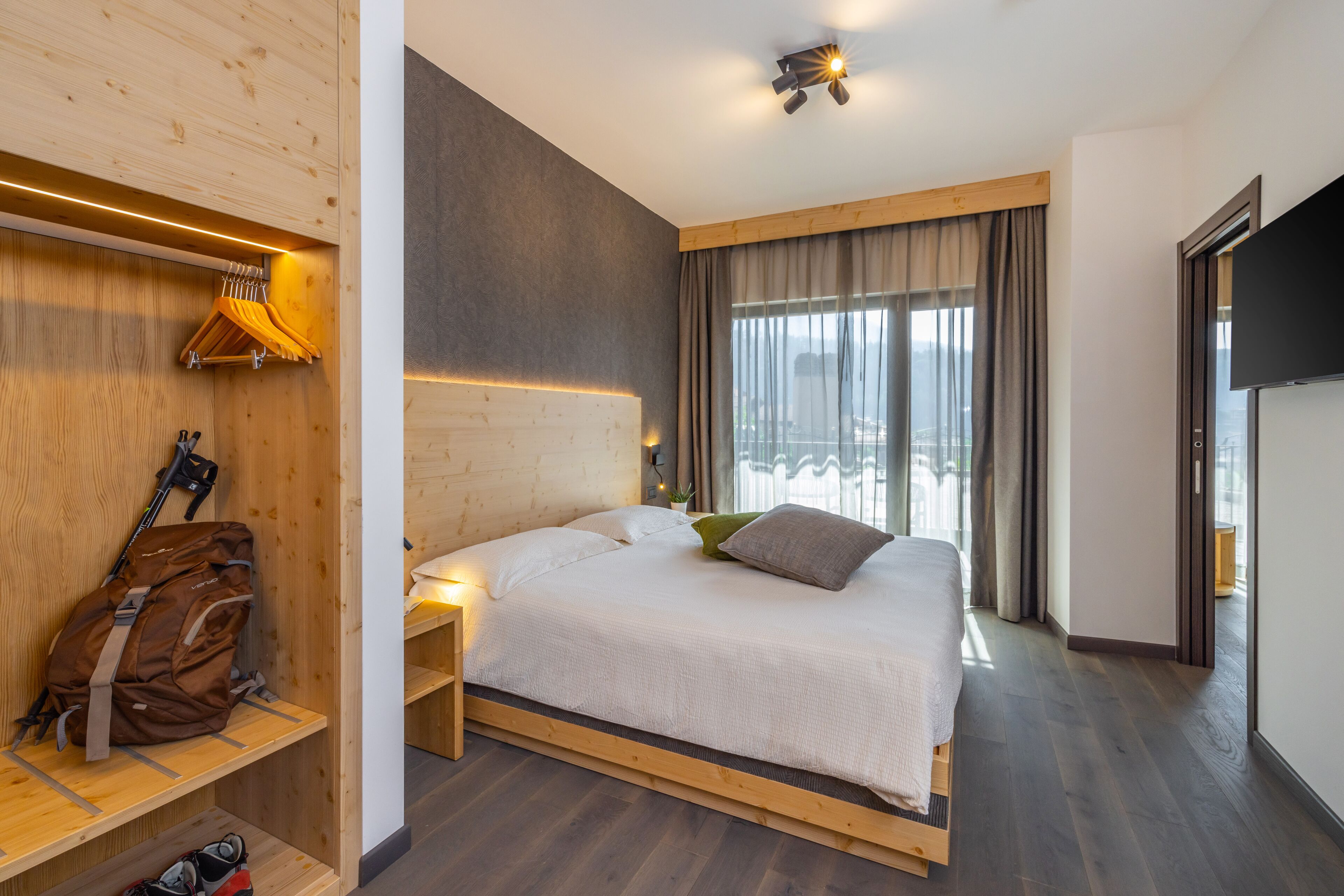 Ein modernes Hotelzimmer mit einem hölzernen Kopfteil, einem großen Bett mit weißer Bettwäsche, einem Nachttisch mit Lampe, einem an der Wand montierten Fernseher und einem Fenster mit Vorhängen.