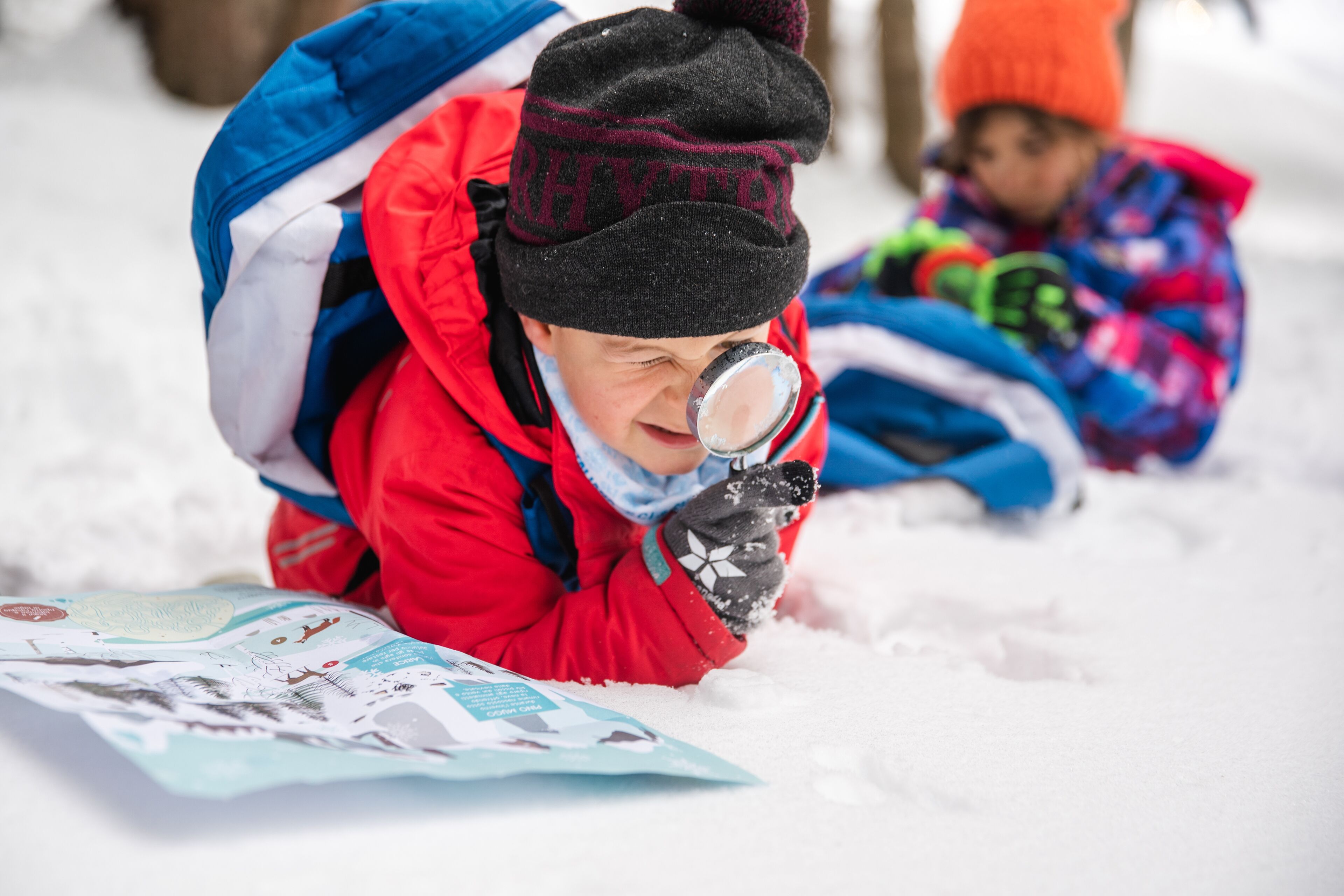 A child in a red jacket and black hat with a magnifying glass, lying on the snow.