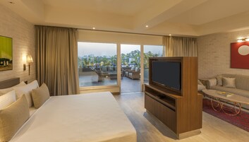 A hotel room with a large bed, a TV, a sofa, and a balcony with a view of the city.