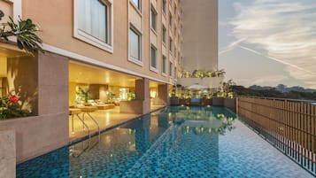 Área de piscina de hotel com piso ladrilhado, espaço de estar com espreguiçadeiras e vista para o horizonte urbano.