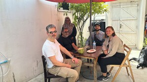 A group of people seated at an outdoor table with a red umbrella, a coconut drink, and a water bottle. - Goby Lodge Guest House (Mahibadhoo)