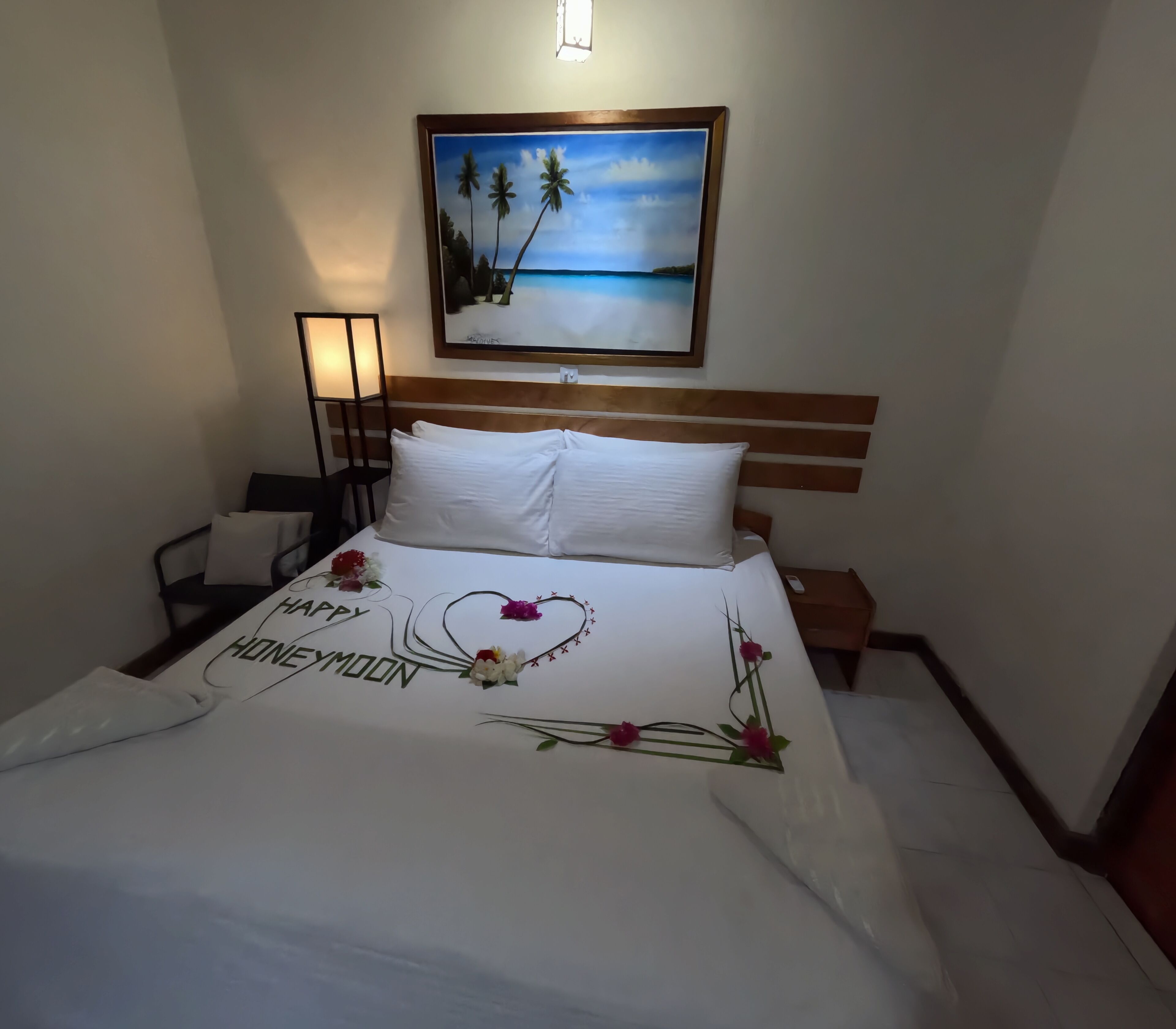 A neatly made bed with a "Happy Honeymoon" message, a painting of a beach scene above it, and a bedside table with a lamp.