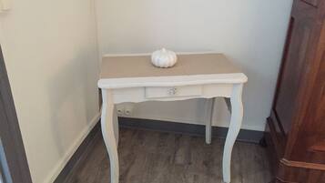 A small white table with a drawer, placed in a corner with a wooden floor and a wooden cabinet in the background.