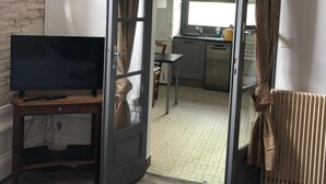 A room with a television on a wooden stand, a door leading to a kitchen with a window, and a radiator. - Villa des T (Arcachon)