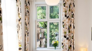 A room with a large window, curtains with a leaf pattern, and a vase with flowers. - Faust and Mona Lisa Apartments (Kraków)