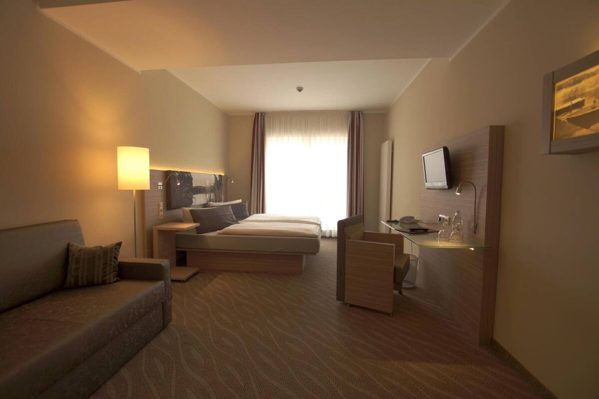 Ein Hotelzimmer mit einer Couch, einem Bett, einem Schreibtisch, einem Fernseher und einem Fenster mit Vorhängen.