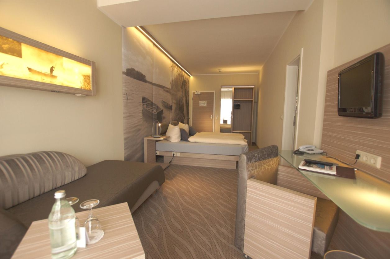 Habitación de hotel compacta con una cama, un sofá, una mesita con una botella y un vaso, un televisor montado en la pared y un cuadro.
