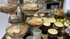 Dispositiu de buffet amb diversos aliments i recipients, incloent bols etiquetats 'Smacks' i 'Cereal Bars', i una varietat de bols i gots sobre una taula de fusta.