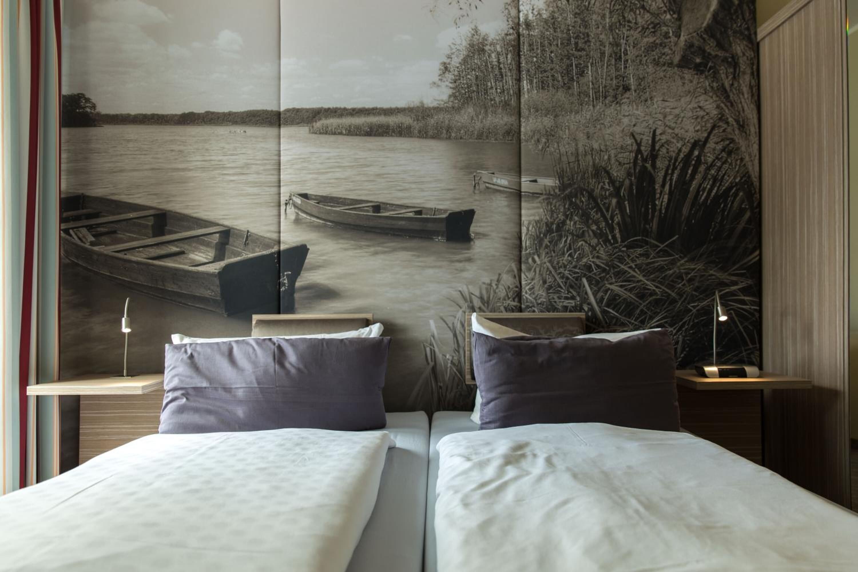 Ein Schlafzimmer mit zwei Betten, einem großen Fenster und Blick auf einen Fluss sowie Booten, und einem Nachttisch mit Lampe.