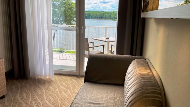 Habitació d'hotel amb un sofà, vista a un balcó amb taula i cadires, i una finestra gran amb cortines.