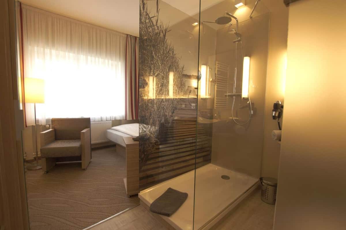 Ein modernes Hotelzimmer mit einer gläsernen Dusche, einem Sofa, einem Bett und Blick auf eine Treppe und einen Außenbereich.