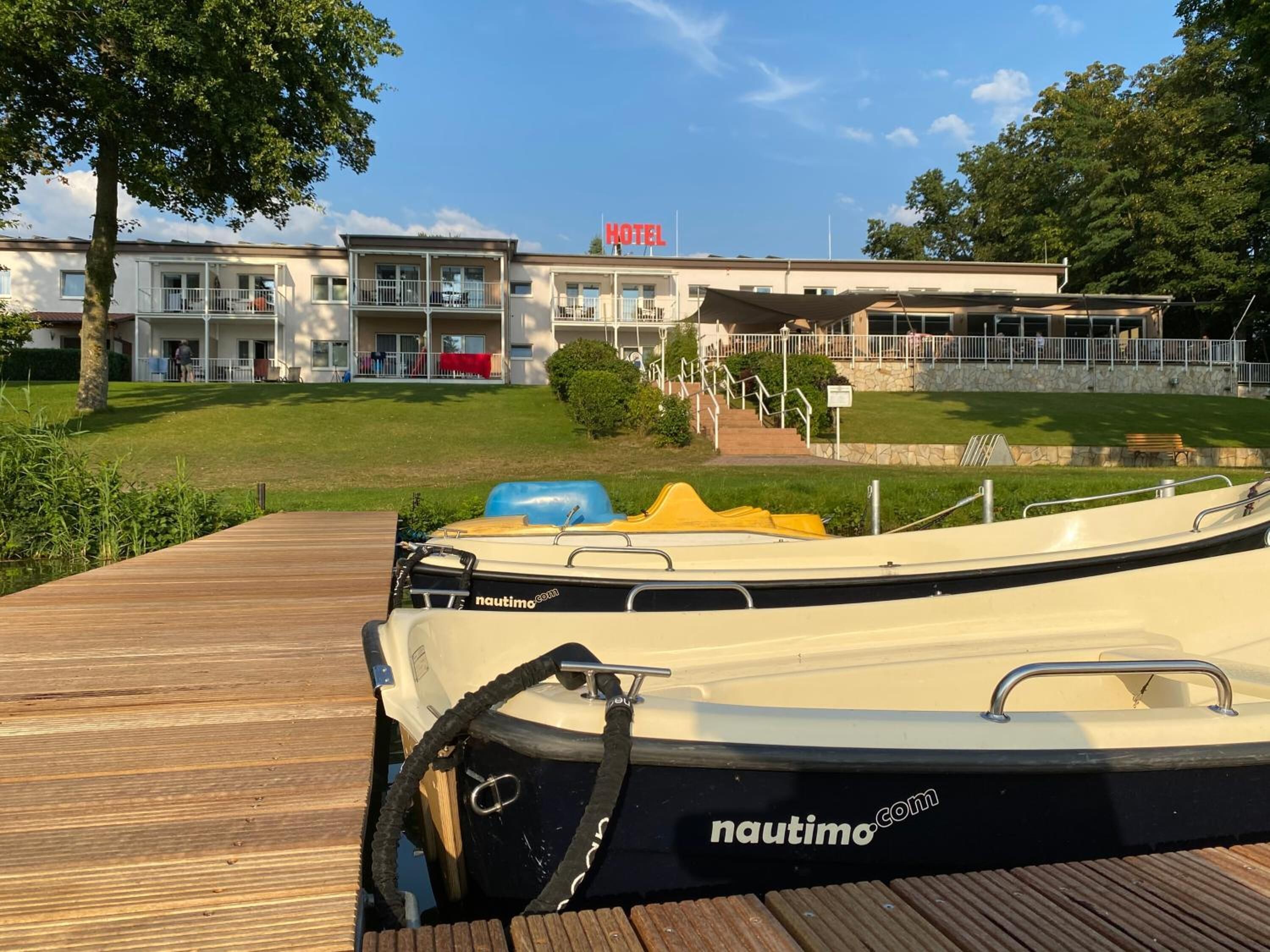 Une jetée en bois avec un bateau blanc et jaune amarré, portant le nom « nautimo.com ». Au fond, on aperçoit un hôtel avec une enseigne indiquant « HÔTEL ».