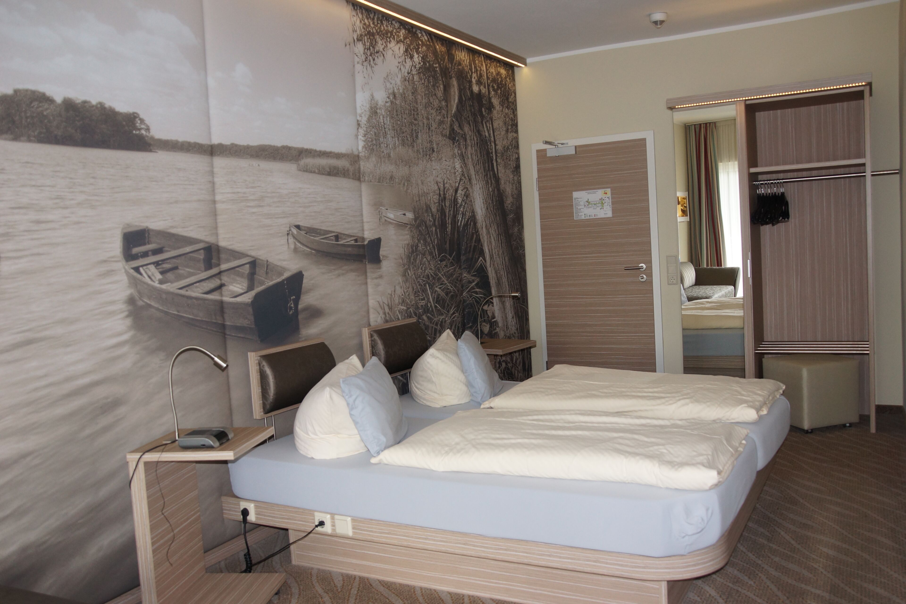 A hotel room with a large wall mural of a lake scene, a bed with white linens, a bedside table with a lamp, and a wardrobe.