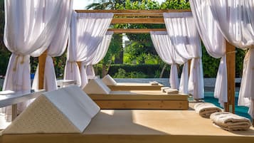 Una pérgola de madera con cortinas blancas, una piscina y tumbonas con toallas.