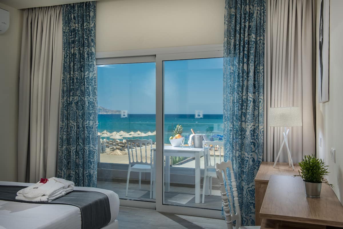 Ett hotellrum med en säng, ett litet bord med en växt och utsikt över stranden och havet genom stora glasdörrar.