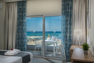Una habitación de hotel con una cama, una mesita con una planta y vistas a la playa y al océano a través de amplios ventanales.