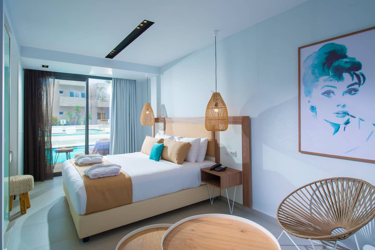 Ett modernt hotellrum med en stor säng, ett litet bord, en flätad stol och utsikt över poolen.
