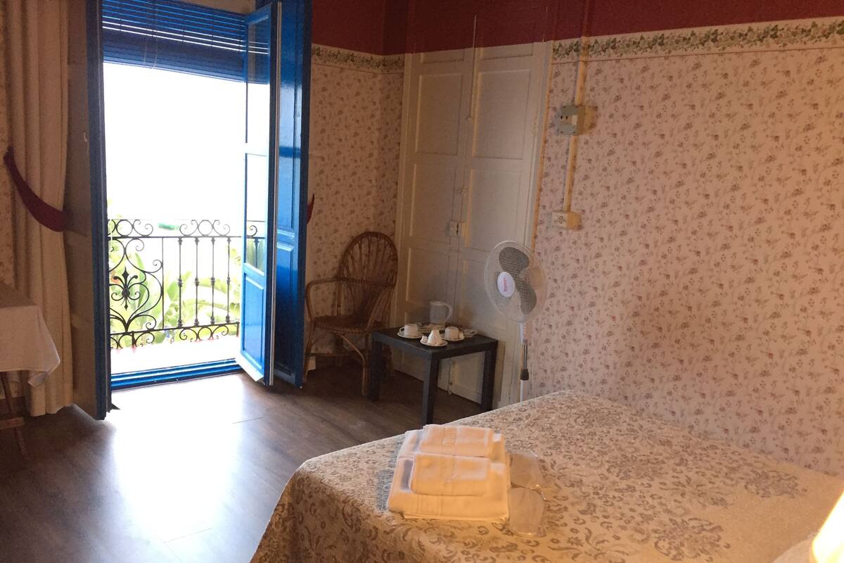 Una habitación con cama, una silla, una mesa con ventilador y una ventana con puerta abierta.