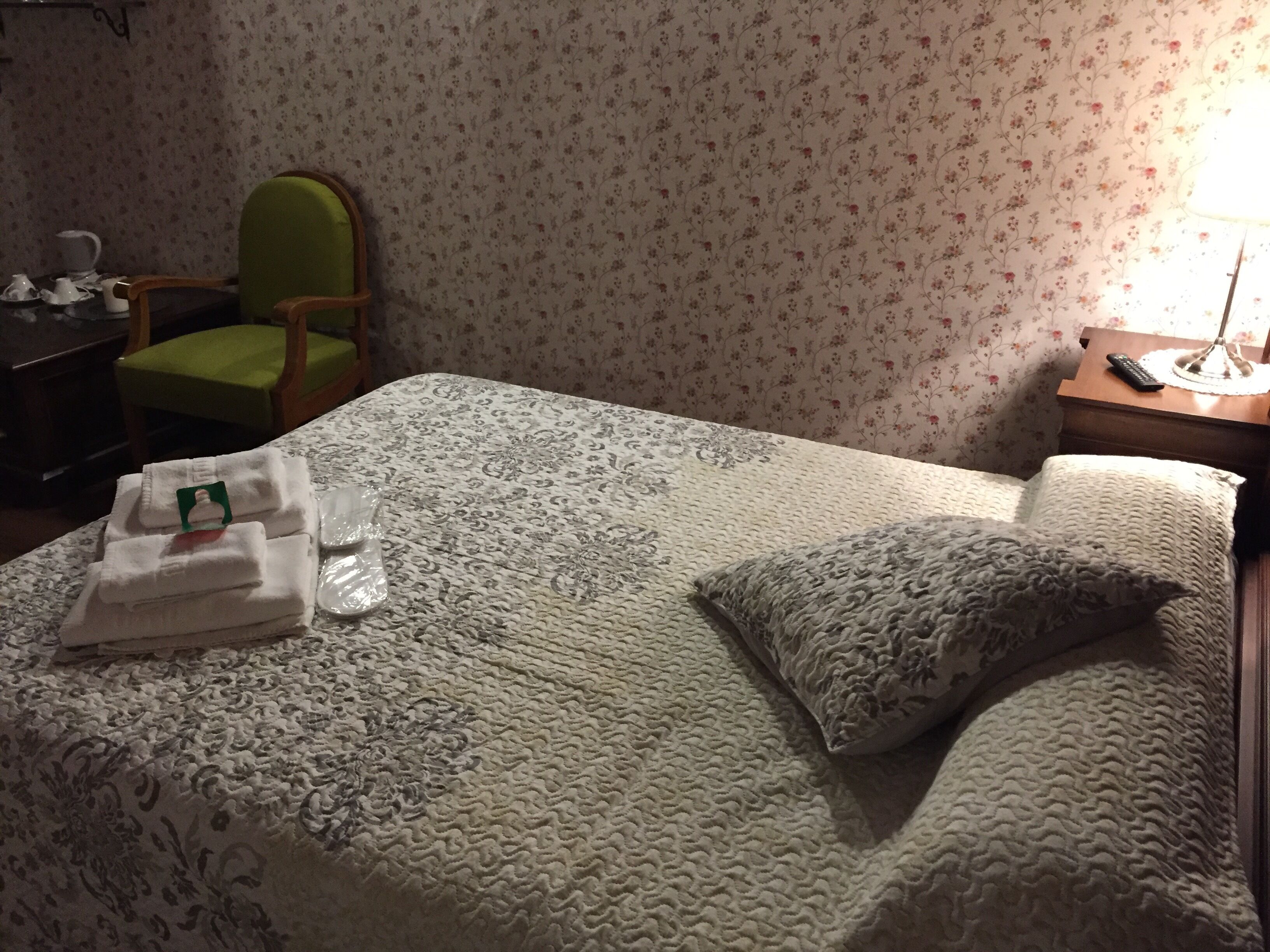 Una cama bien tendida con una colcha estampada, una silla verde, una mesita con tetera y una mesita de noche con lámpara.