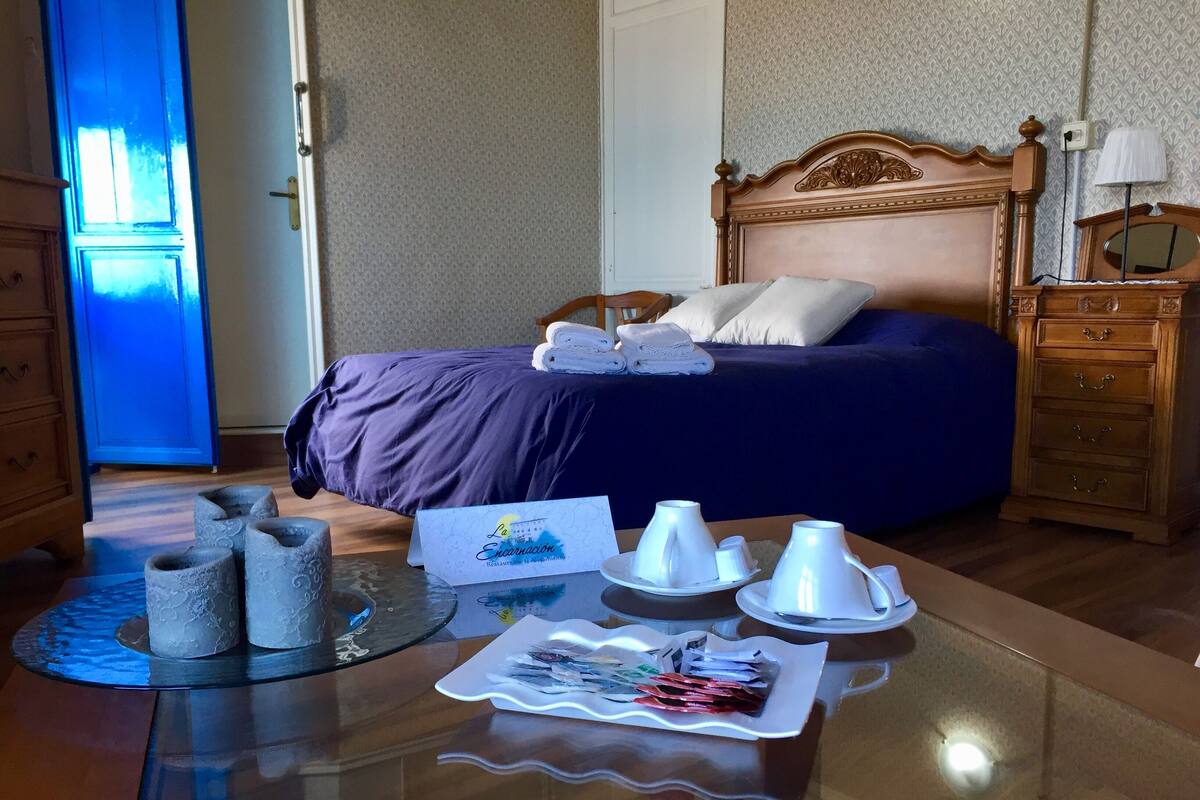 Habitación de hotel con una cama, una mesita de noche de madera, una mesa de vidrio con artículos de tocador y una puerta azul.