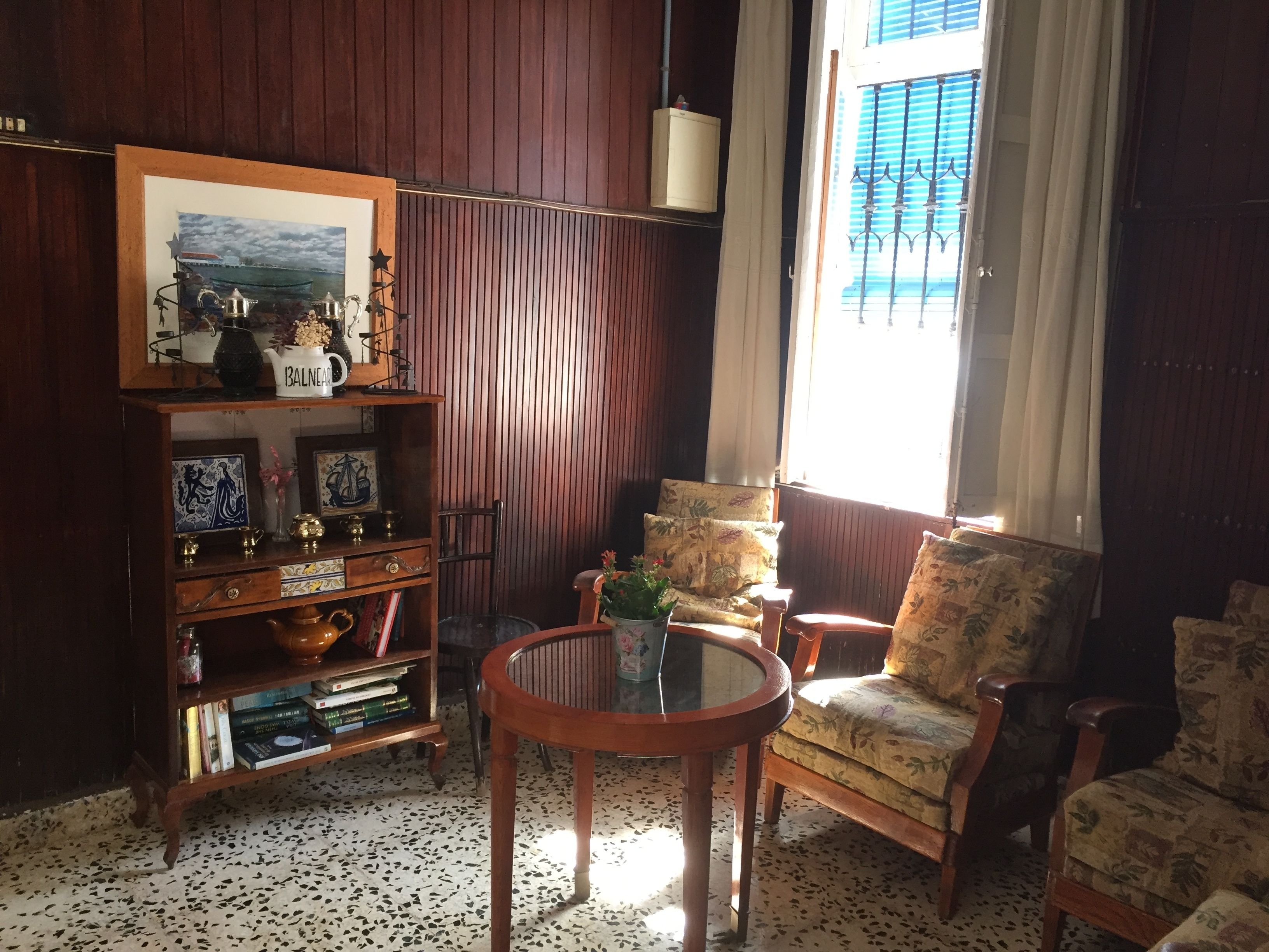 Una habitación acogedora con paneles de madera, una estantería con objetos decorativos, una mesa redonda y dos sillones con estampado.