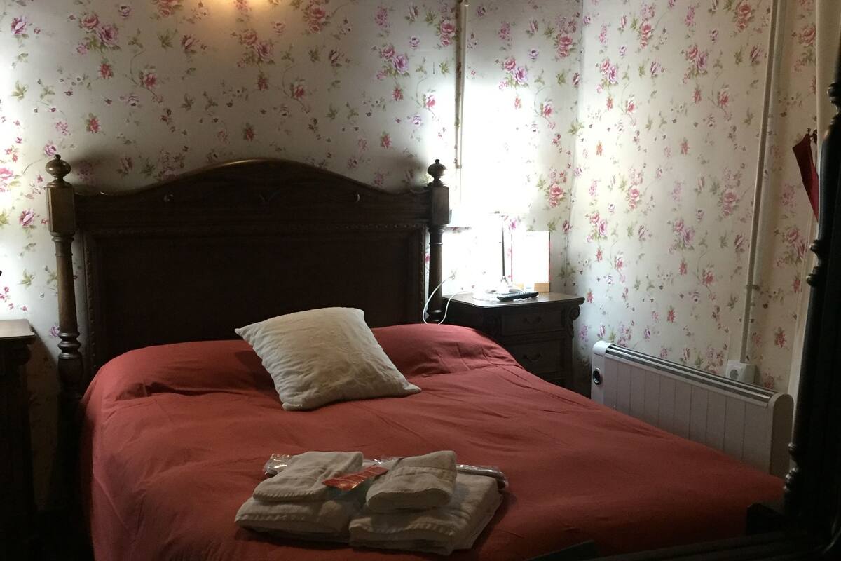 Un dormitorio con cama de madera, papel tapiz floral, una mesita de noche y una lámpara de noche.