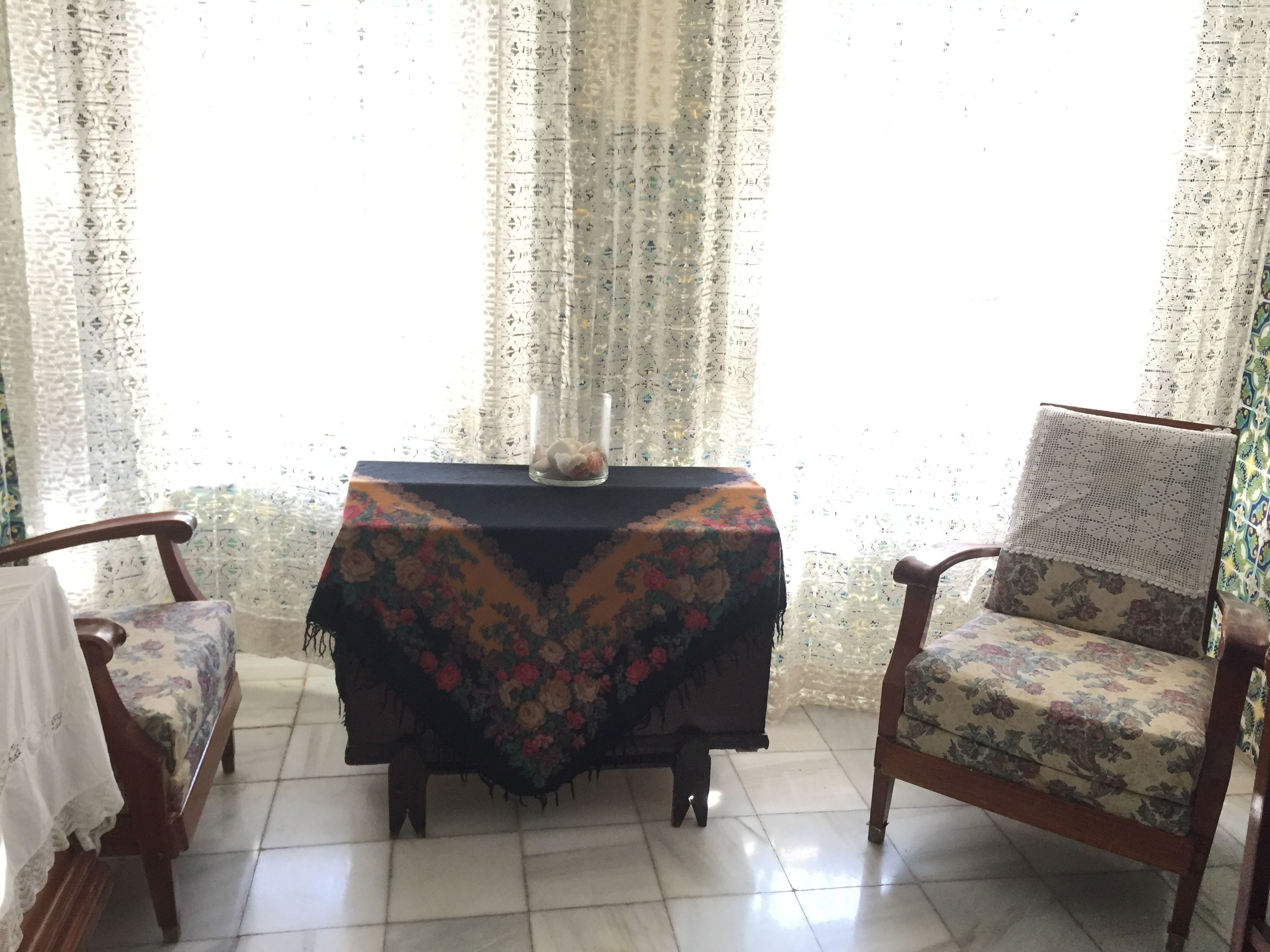 Una habitación con un sofá estampado con flores, una silla de madera, una mesita con un jarrón y una ventana con cortinas traslúcidas.
