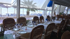 Un espace de restauration en plein air, avec des chaises et des tables en osier disposées pour un repas, donnant sur une plage parsemée de parasols et de chaises.