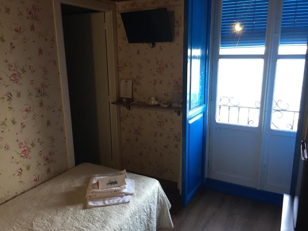 Un dormitorio con papel tapiz floral, una cama con toallas dobladas, una estantería con cafetera y una ventana con vistas al exterior.