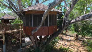 Ein Baumhaus mit einem Reetdach, umgeben von Bäumen und einer Holzterrasse.