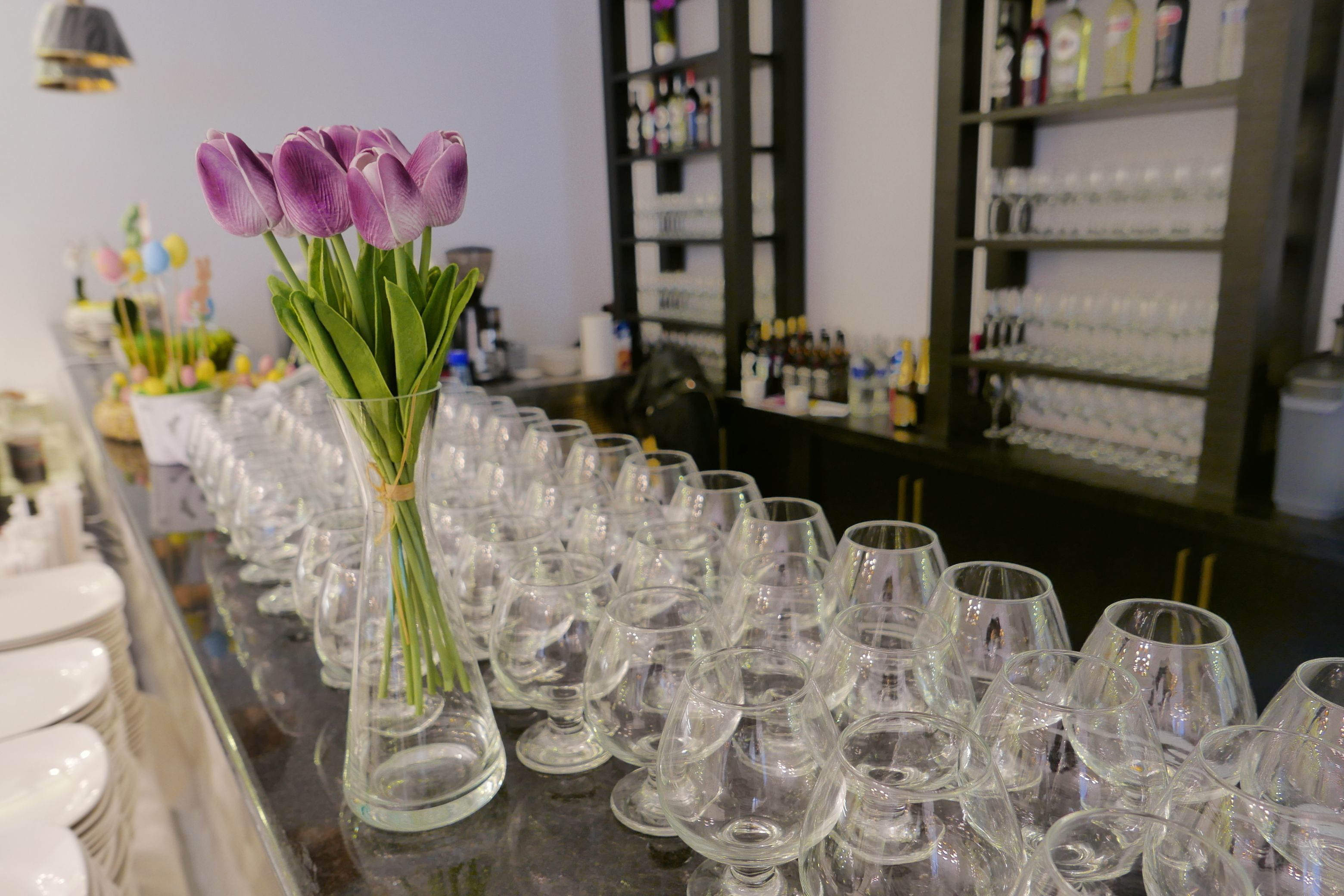 Una zona de bar con un jarrón de vidrio que contiene tulipanes morados y numerosos vasos de vino vacíos alineados sobre la barra.