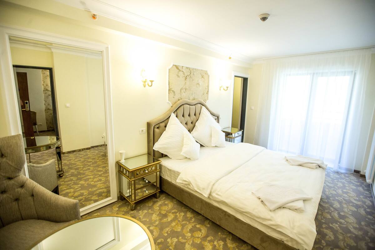 A hotel room with a large bed, a sitting area with a chair, a nightstand, and a mirror.